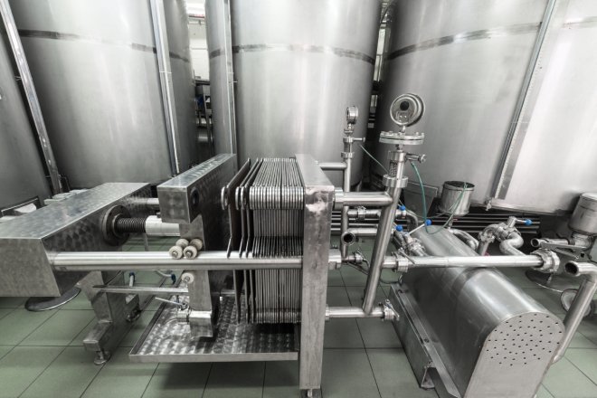 A large stainless steel automatic filter press sits in a room with large stainless steel storage containers.