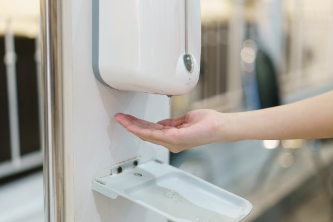 A touchless soap dispenser attached to a bathroom wall next to a mirror. A cupped hand is reaching out underneath it.