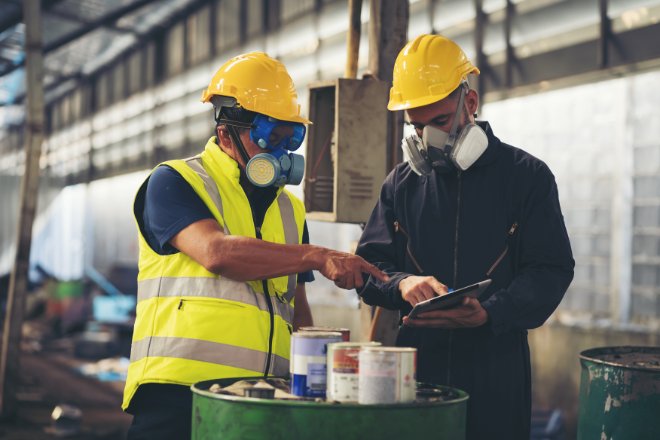 Two workers wearing hard hats and respirators review a tablet while inspecting equipment in an industrial facility.