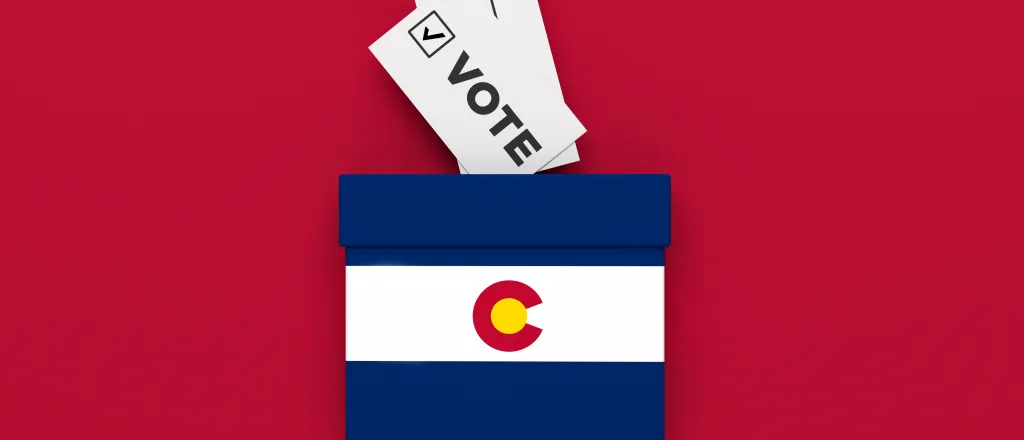 PROMO HIRES Election Colorado Vote Ballot - Baris-Ozer - iStock-1420488382 Miniature ballot box with and image of the Colorado state flag on a red background with slips of paper representing ballots