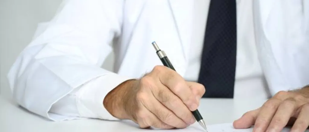 man in a medical coat, button down shirt, and tie writing on a piece of paper with a black mechanical pencil