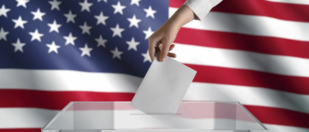 Hand placing a piece of pager into a clear ballot box in front of the United States flag