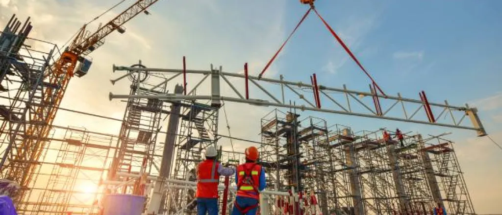 Two construction workers looking up as a steel truss is lifted by a large crane while the sun sets in the background.