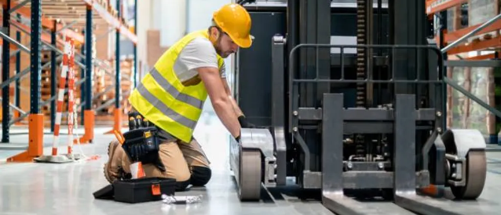 A mechanic kneeling next to a forklift in a warehouse as he makes repairs with a tool kit by his side. A mechanic kneeling next to a forklift in a warehouse as he makes repairs with a tool kit by his side.