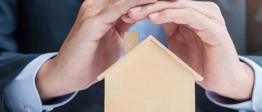 Safety features your rental property can’t go without A man in a suit sitting at a table and covering the roof of a small wooden home model with both hands.