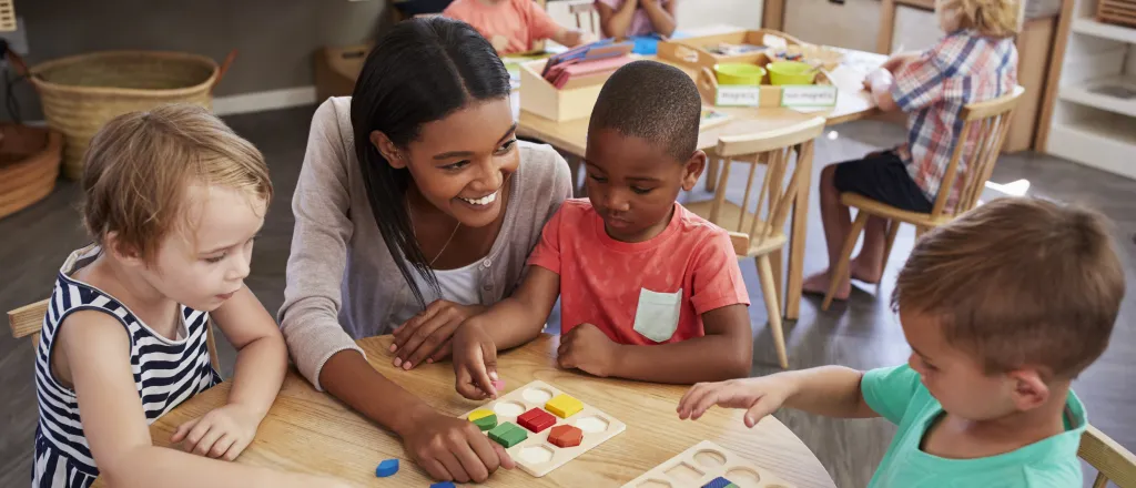 PROMO HIRES Education - Childcare Children School - monkeybusinessimages - iStock-684060608 Young children and a care giver at tables engaged in educational activities.