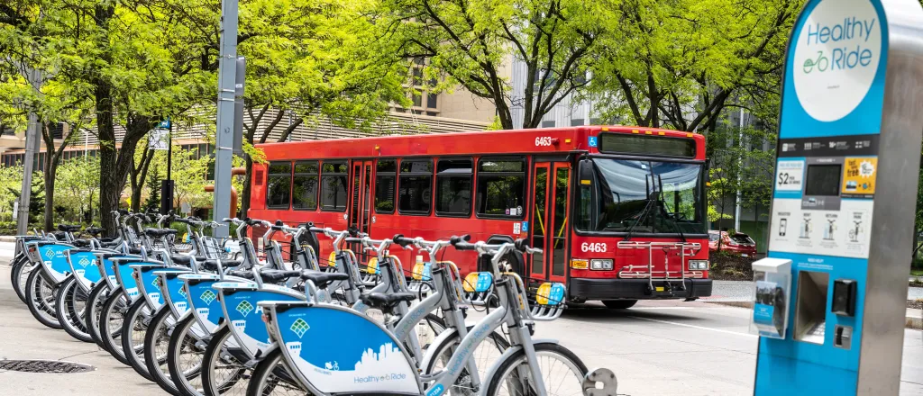 PROMO HIRES Transportation - Public Transit Bike Bicycle Bus - arlutz73 - iStock-1323085178 Row of electric bicycles for rent with a passenger bus in the background.