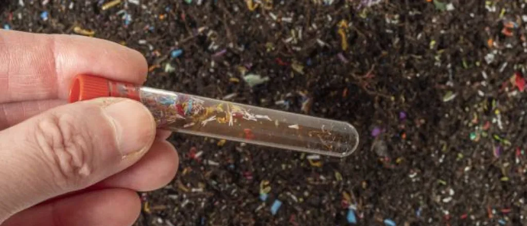 A hand holding a test tube containing a soil sample above a patch of dirt littered with colorful microplastic shards. A hand holding a test tube containing a soil sample above a patch of dirt littered with colorful microplastic shards.