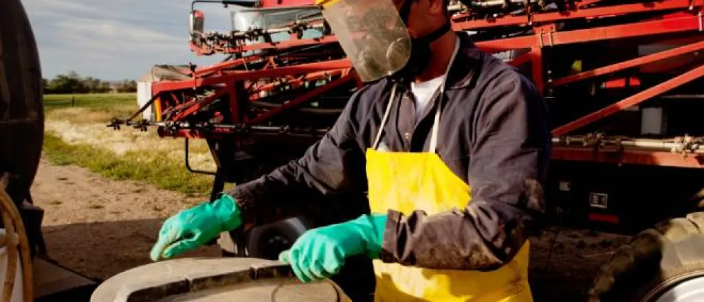 A person wearing a protective shield, a yellow protective apron, green protective gloves, and a mask is opening a container. A person wearing a protective shield, a yellow protective apron, green protective gloves, and a mask is opening a container.