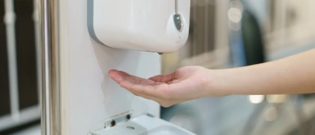 A touchless soap dispenser attached to a bathroom wall next to a mirror. A cupped hand is reaching out underneath it.