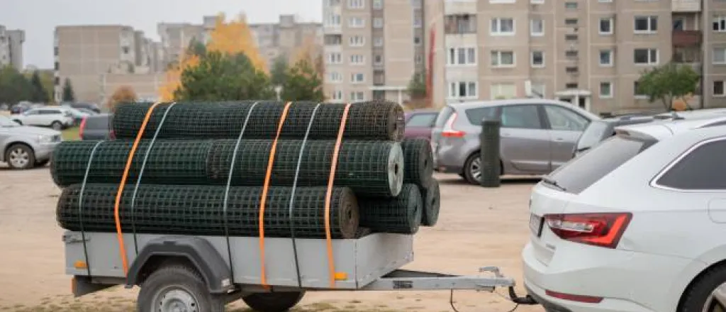 A parked white car with a trailer hitch and a utility trailer behind it carrying rolls of metal fencing strapped to the trailer. A parked white car with a trailer hitch and a utility trailer behind it carrying rolls of metal fencing strapped to the trailer.