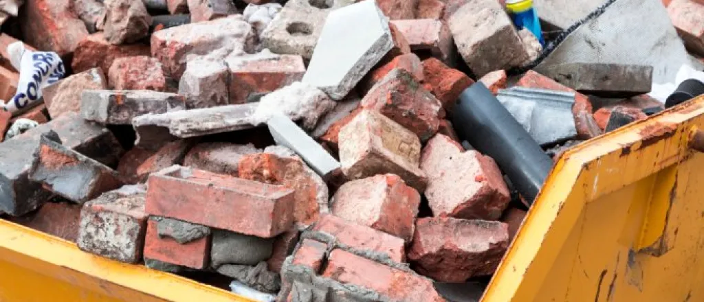 Close-up of a yellow dump truck bed. The bed is full of bricks of various sizes and colors, as well as other debris.