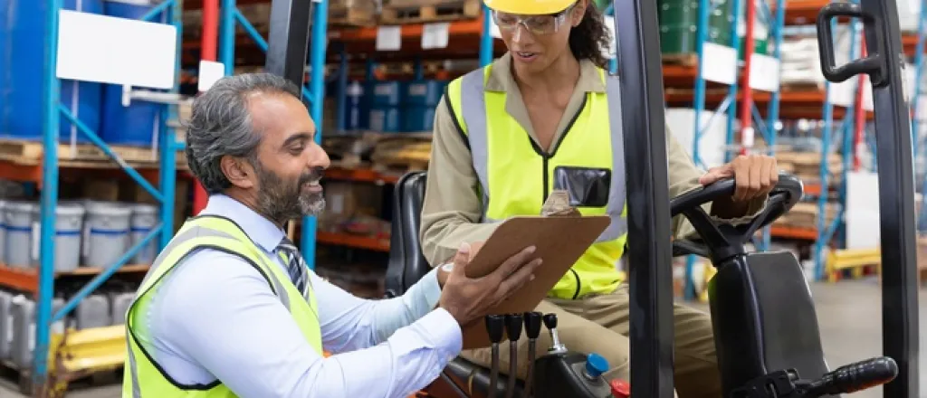 The most common warehouse OSHA violations Two workers wearing safety vests. One is sitting in a forklift while the other is showing them information on a clipboard.