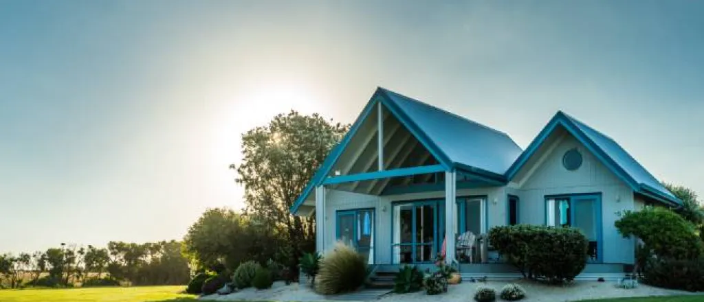 A luxurious home sits on a wide and empty plot of land. There is greenery around the home and the sun behind it.