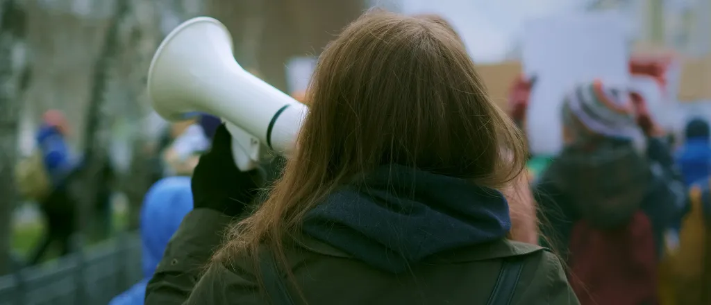 PROMO HIRES CROPPED Miscellaneous - Protest Demonstration Politics Community - sibway - iStock-1766290065 View from behind of a person holding a megaphone at a protest rally.