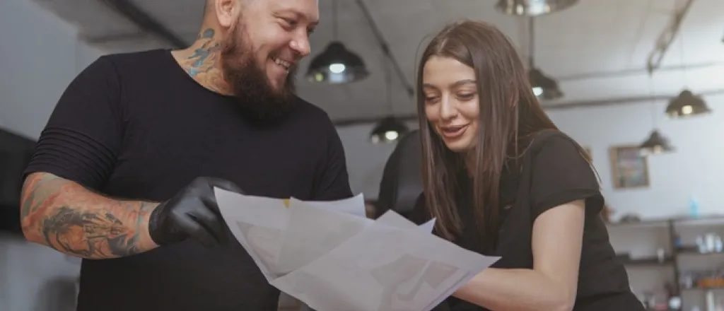 A tattoo artist wearing black latex gloves showing a smiling client several printed images for them to consider. A tattoo artist wearing black latex gloves showing a smiling client several printed images for them to consider.