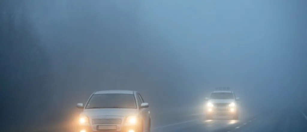 Two white vehicles with their headlights on drive down a straight road at night through a dense fog.