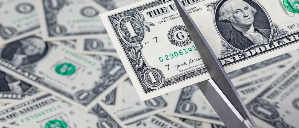 PROMO HIRES Finance - Budget Cut Money Currency Scissors - JJ Gouin - iStock-1642293566 A pair of scissors is preparing to cut a United States one dollar note. In the background, additional dollar bills are strewn haphazardly.