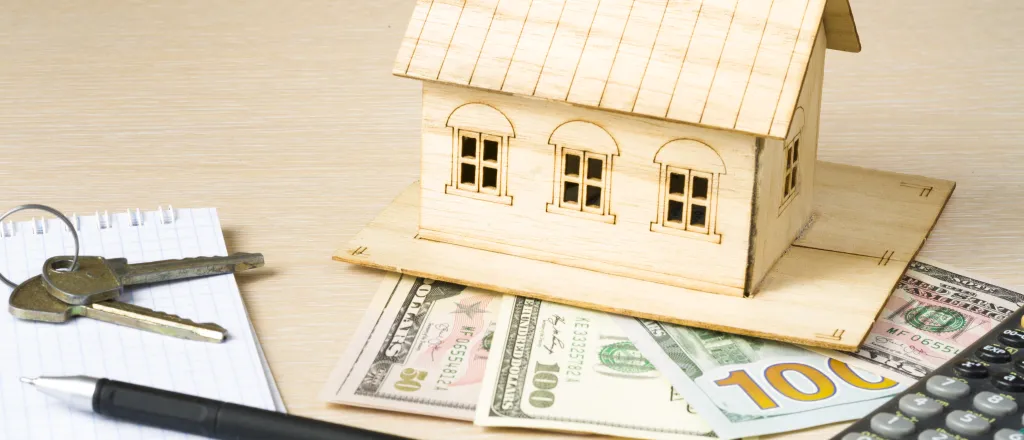 PROMO HIRES Finance - Money House Calculator Keys Pen Paper - iStock - Motizova Model house made of wood resting on United State currency. A calculator, pen, notepad, and keys are nearby.