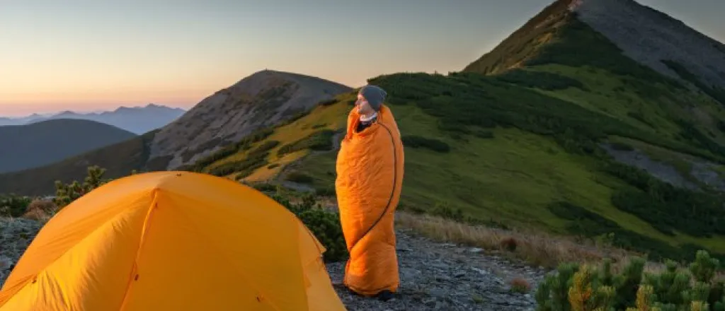 A man wraps himself in his orange sleeping bag as he gazes at the mountainous landscape beside his tent.