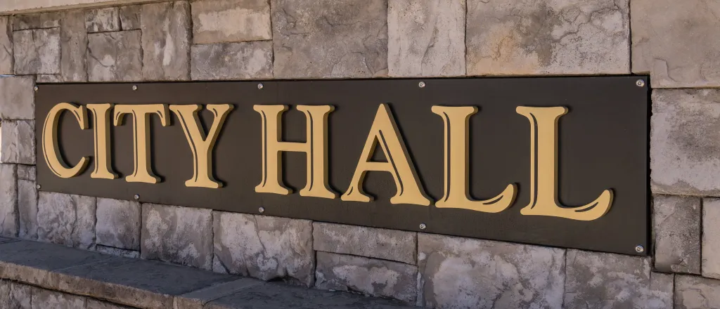 PROMO HIRES Government - Building City Hall Politics - HABesen - iStock-1398867931 Close up of a cut stone wall. A sign affixed to the wall had the words "City Hall"