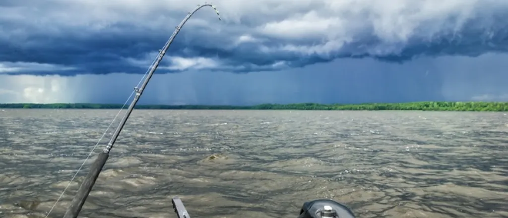 The stern of a boat on a lake before a storm hits. A fishing rod is cast into the choppy water below a dark, cloudy sky.