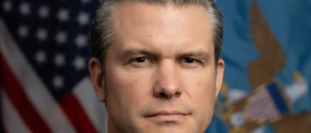 Head shot of Pete Hegseth, Secretary of Defense under the second Trump administration, in front of the flag of the United States and a military flag.