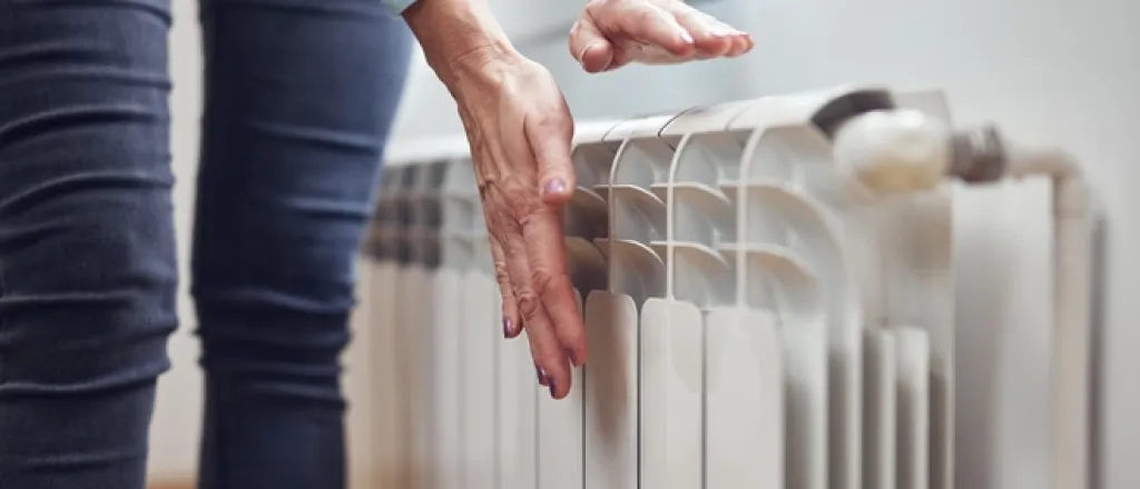 A homeowner in jeans stands at a room's radiator as they place their hands near the vents to feel for heat.
