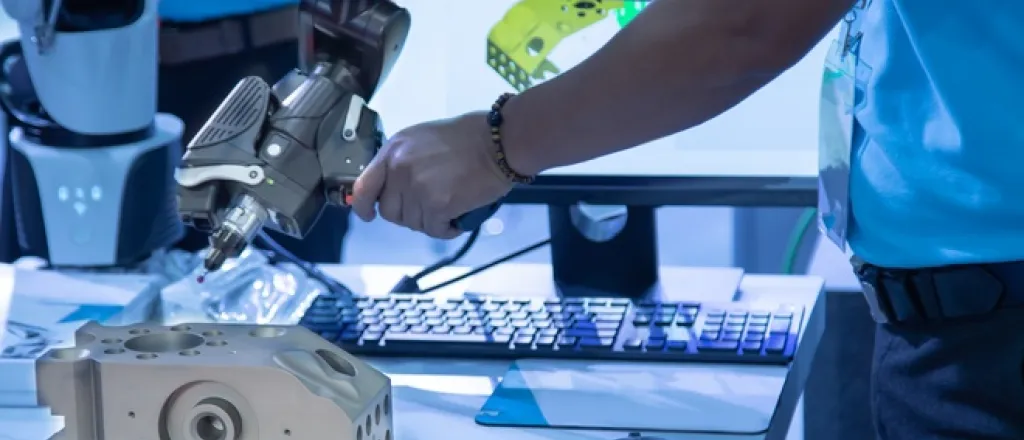 A technician stands in front of a portable 3D measuring arm. A part with grooves and holes rests on the table below the tool.