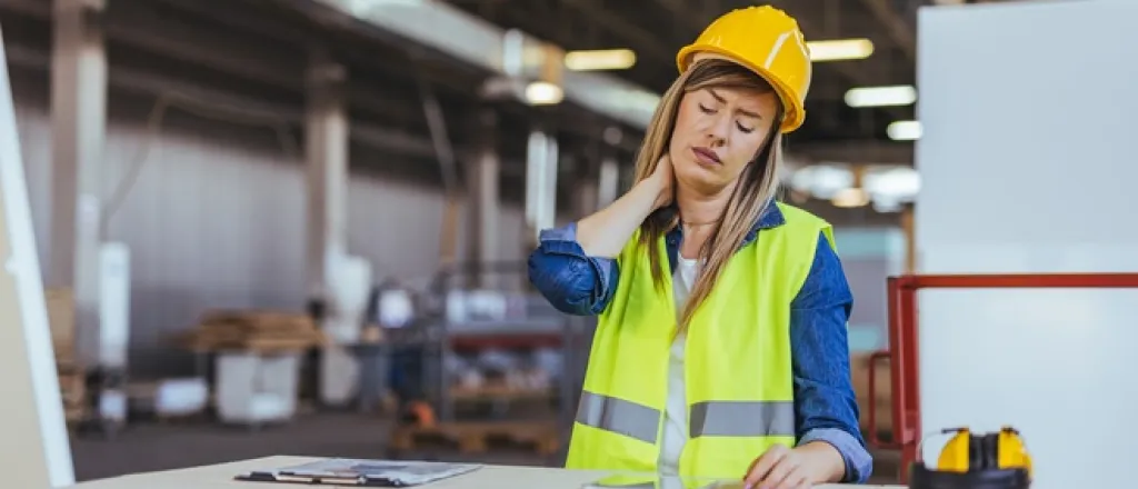 A female engineer sits at a work bench while she holds her neck in pain. She wears a hard hat and a safety vest.