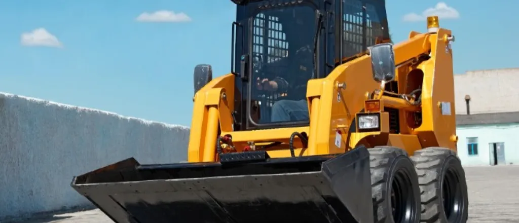 A yellow skid steer loader with a large black front bucket parked on a paved lot under a clear blue sky.