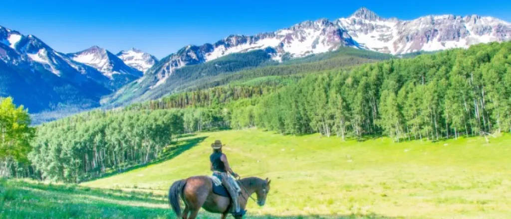 Cowboy riding a horse through a Colorado mountain landscape with grassy hills and alpine scenery in view.