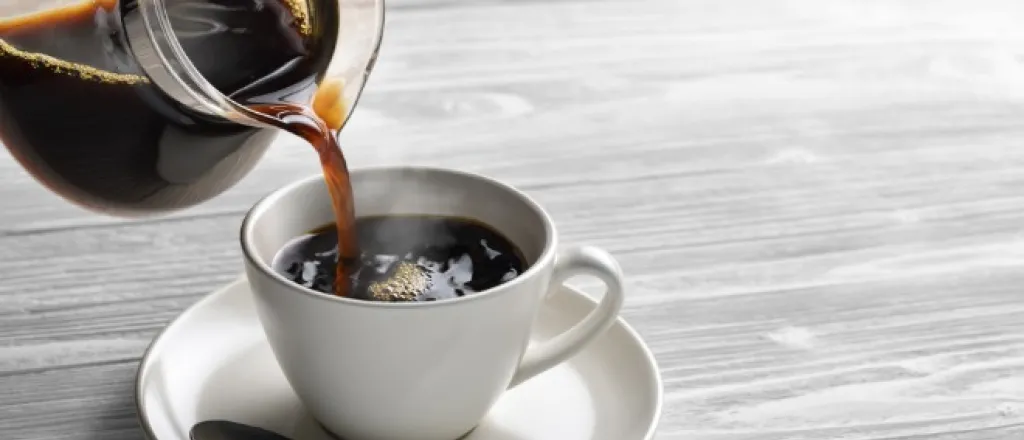 A close-up of a glass coffee pot pouring hot coffee into a white mug on a saucer. A silver spoon rests on the saucer.
