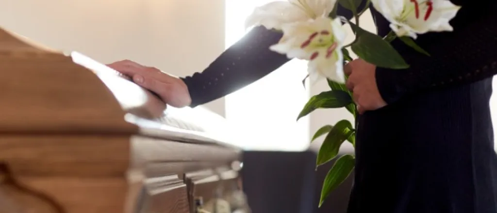 A woman stands in a black dress at a funeral. She holds white lilies in one hand and puts her other hand on the coffin.
