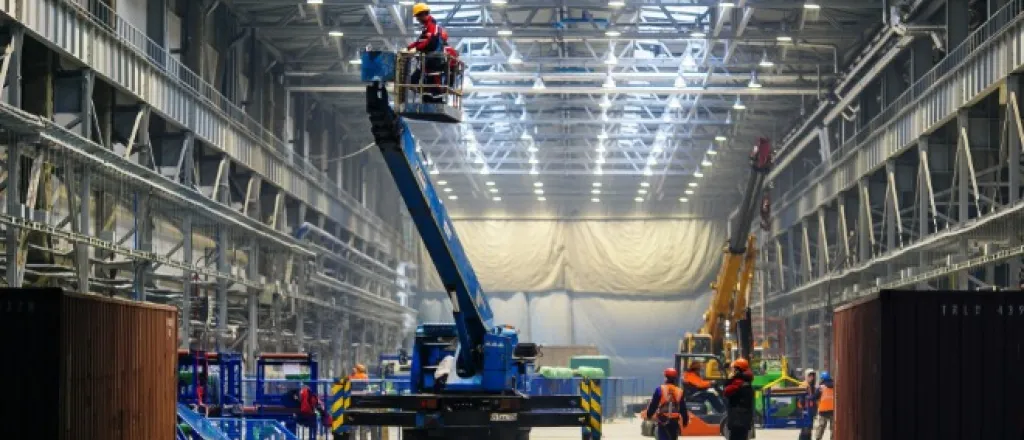 Several people are working in a large industrial space with visible equipment and material around them.