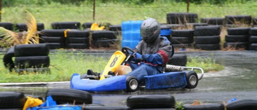 A driver navigates a tight corner of a go-kart track in the rain, with tires forming a safety barrier around them.