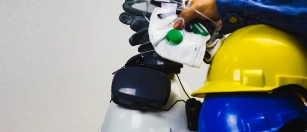 A stack of PPE equipment resting on a table. Someone holds a face mask, safety glasses, and gloves on top of some hard hats.