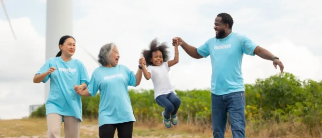 Three volunteers in blue shirts swing a laughing child by the hands on a path near a rural wind turbine and field.