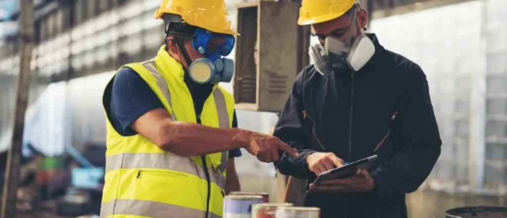 Two workers wearing hard hats and respirators review a tablet while inspecting equipment in an industrial facility.
