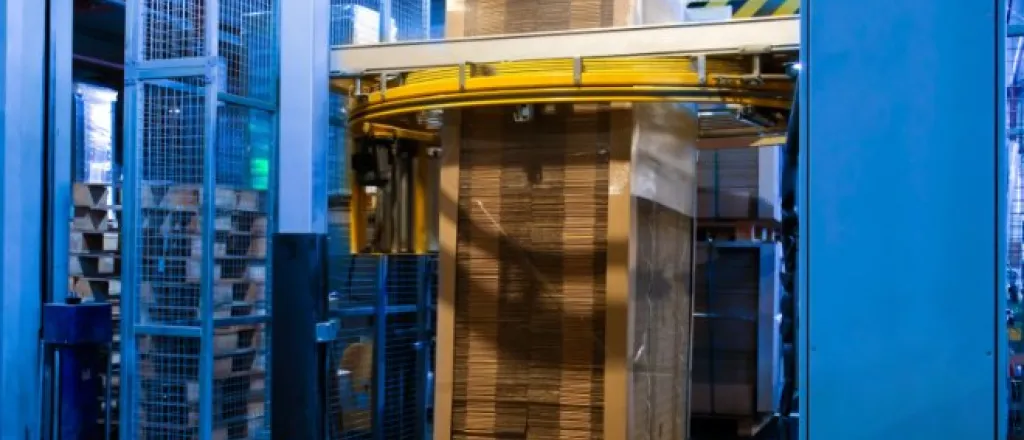 A blue stretch wrapping machine in the middle of applying stretch wrap film to a tall pallet of cardboard boxes.