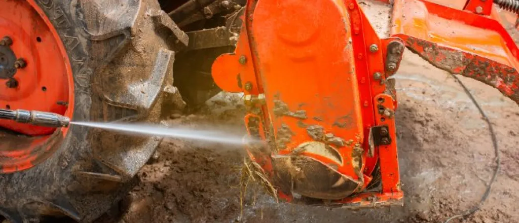 A pressure washer sprays the surface of a muddy red tractor attachment beside a tire. Water pools on the ground.