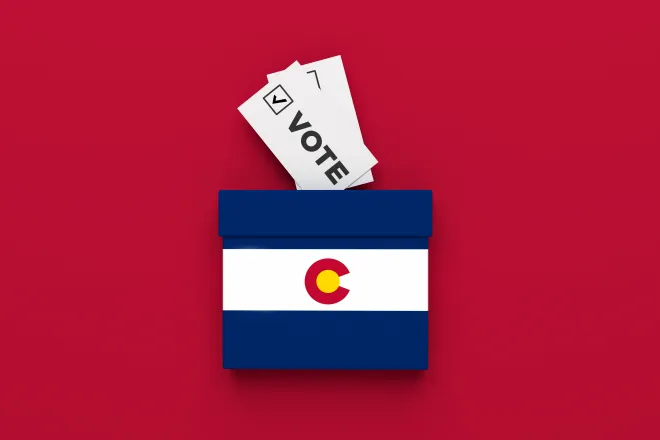 Miniature ballot box with and image of the Colorado state flag on a red background with slips of paper representing ballots