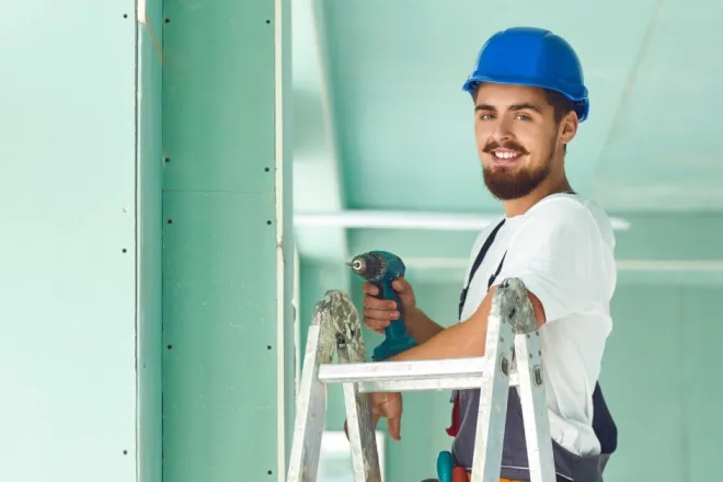 A male worker with a beard and mustache stands at the top of a ladder using a drill to install drywall sheets.