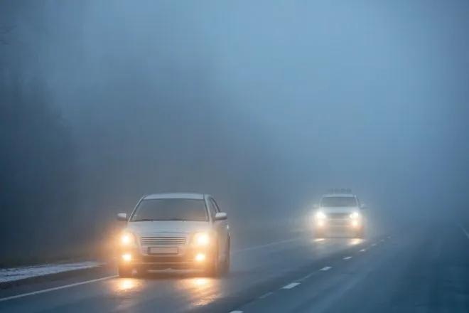 Two white vehicles with their headlights on drive down a straight road at night through a dense fog.