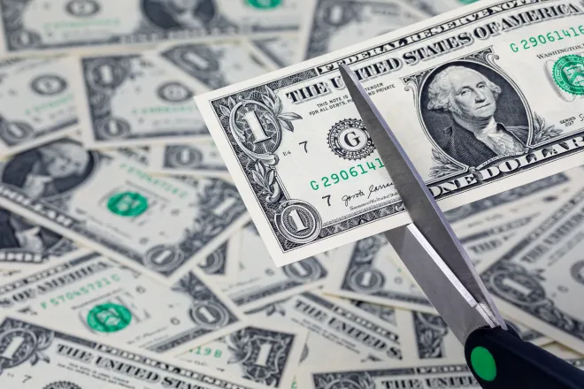 A pair of scissors is preparing to cut a United States one dollar note. In the background, additional dollar bills are strewn haphazardly.