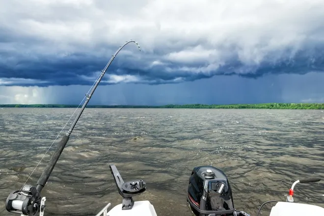The stern of a boat on a lake before a storm hits. A fishing rod is cast into the choppy water below a dark, cloudy sky.