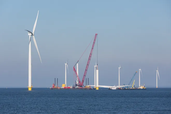 Wind turbines under construction in the ocean. A crane ship is in place to lift a blade assembly.