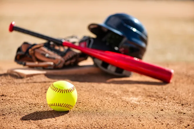 A softball sit in the foreground, with a helmet, bat, and glove sitting on a base slightly blurred in the background.