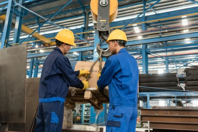 Two people are handling a box that is attached to a pully system. The people are both wearing blue coveralls.