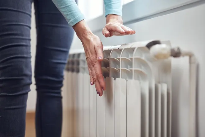 A homeowner in jeans stands at a room's radiator as they place their hands near the vents to feel for heat.